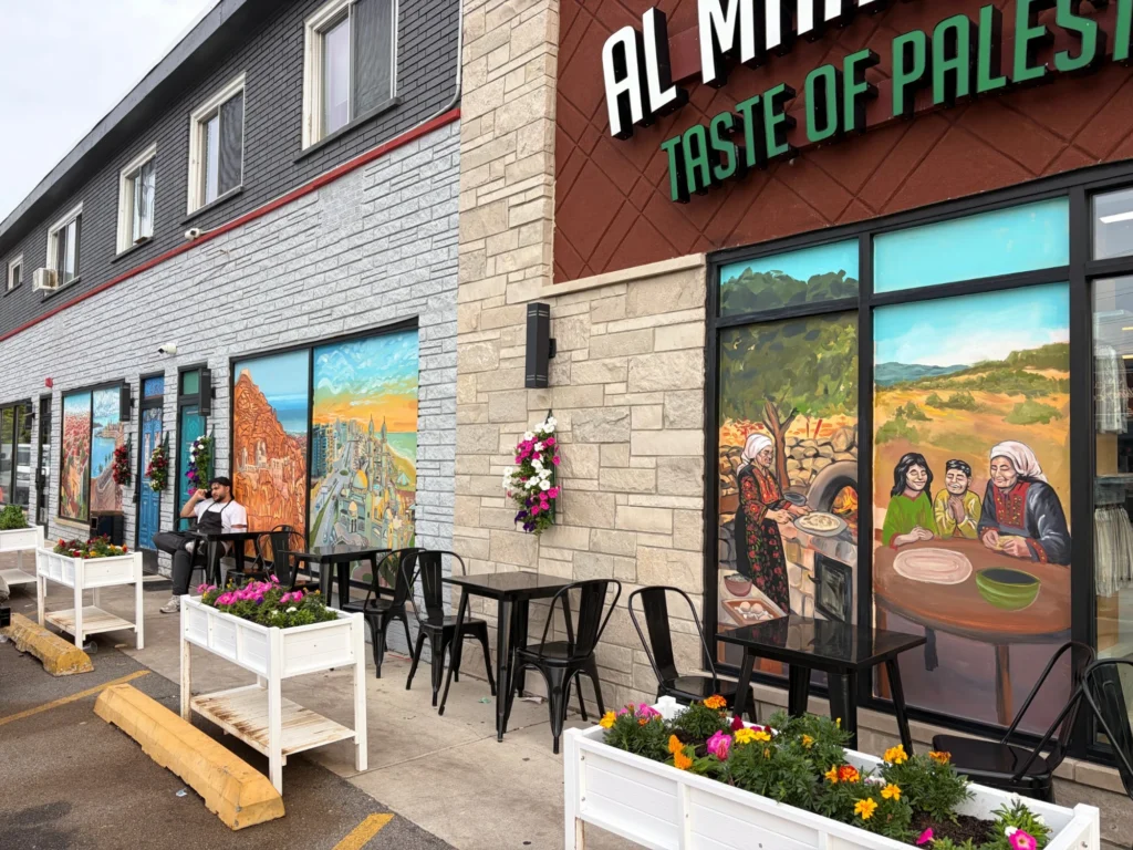 Al Manakeesh Storefront Murals by Caleb Wassell and Abid Shalabi: painted windows along the Al Manakeesh Taste of Palestine storefront showing Nazareth, Akka, Jericho, Gaza City and a grandmother baking bread at a taboon.