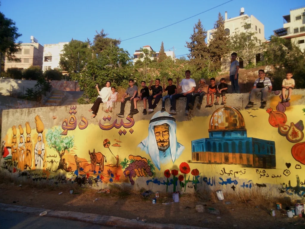 Beit Duqqu Mural, mural by Caleb Wassell: the finished community wall in Beit Duqqu, Palestine with the village name in Arabic, women carrying jars, an elder in a keffiyeh, the Dome of the Rock and red poppies, with the painters seated on top.
