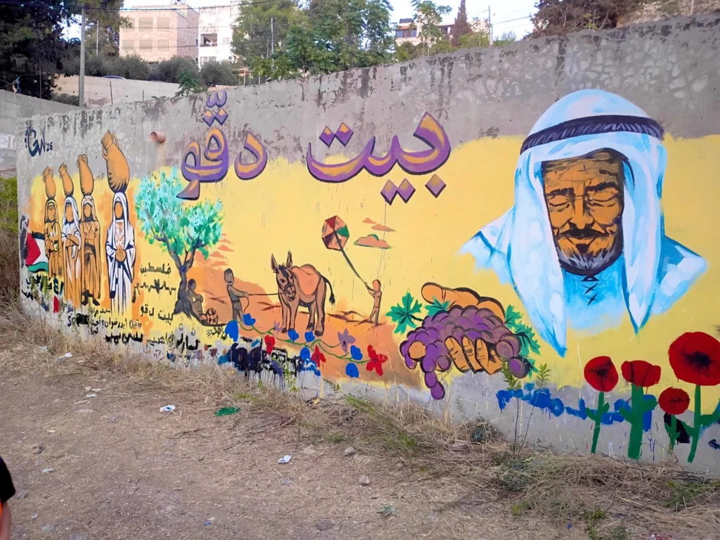 Beit Duqqu Mural, Detail, mural by Caleb Wassell: women carrying clay jars, an olive tree, children with a donkey and kite, a hand holding grapes, an elder in a keffiyeh and red poppies beneath the village name in Arabic in Beit Duqqu, Palestine.