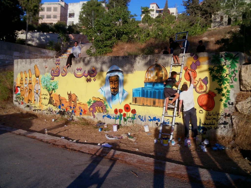 Beit Duqqu Mural, In Progress, mural by Caleb Wassell: community members paint a wall in Beit Duqqu, Palestine with women carrying clay jars, an olive tree, an elder in a keffiyeh, the Dome of the Rock, poppies, figs and pomegranates.