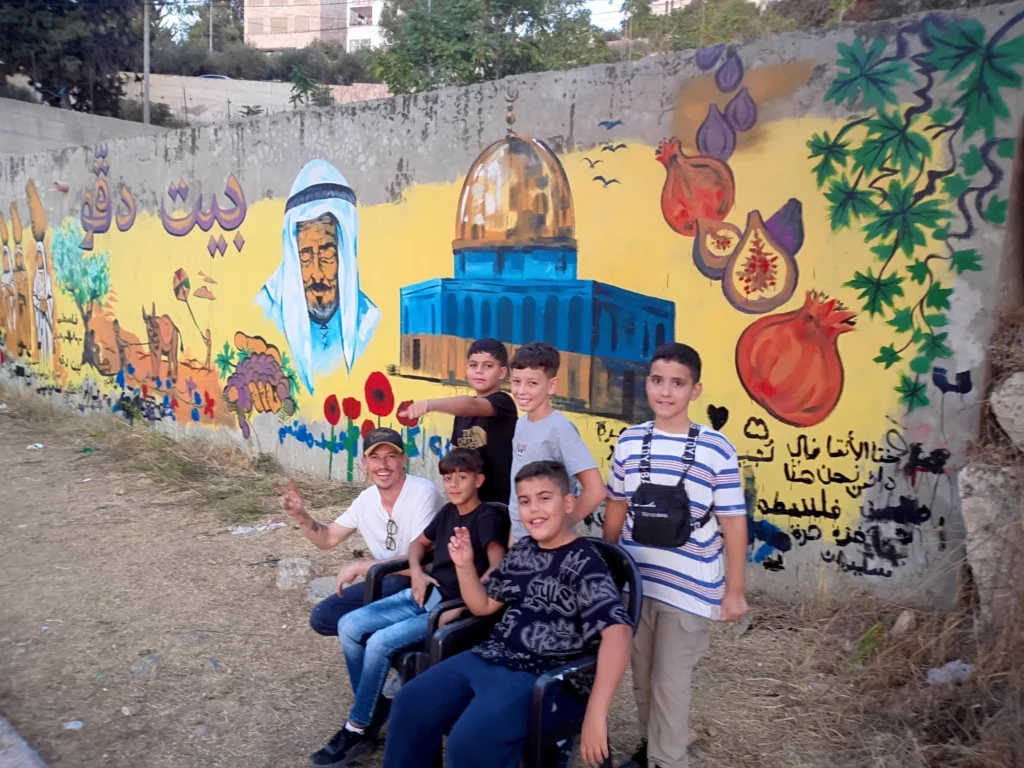 Beit Duqqu Mural, With the Young Painters: Caleb Wassell with the young painters in front of the finished Beit Duqqu mural in Palestine, with the Dome of the Rock, an elder in a keffiyeh, figs and pomegranates.