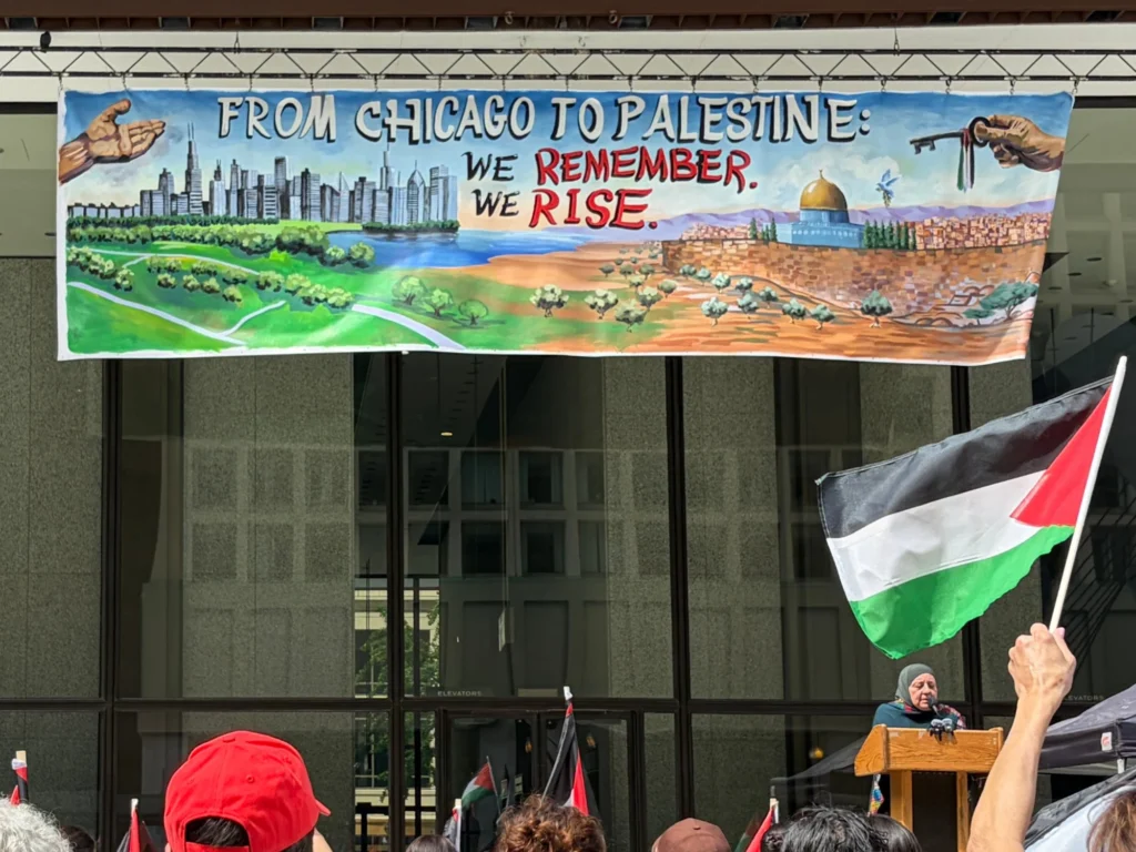 From Chicago to Palestine: We Remember. We Rise., banner by Caleb Wassell: the Chicago skyline and lakefront flowing into olive groves and Jerusalem with the Dome of the Rock, hung above a rally stage.