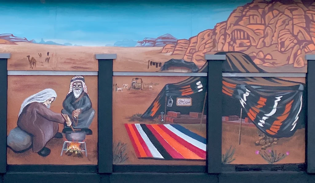 Gaza on My Mind, Bedouin Camp, mural by Caleb Wassell: an elderly couple cooking over a fire beside a striped rug and a black Bedouin tent below red desert cliffs.