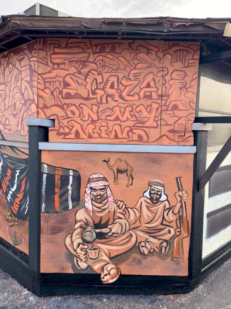 Gaza on My Mind, Desert Coffee, mural by Caleb Wassell: two Bedouin men in keffiyehs share Arabic coffee beside a tent and camel beneath the lettering Gaza on My Mind.