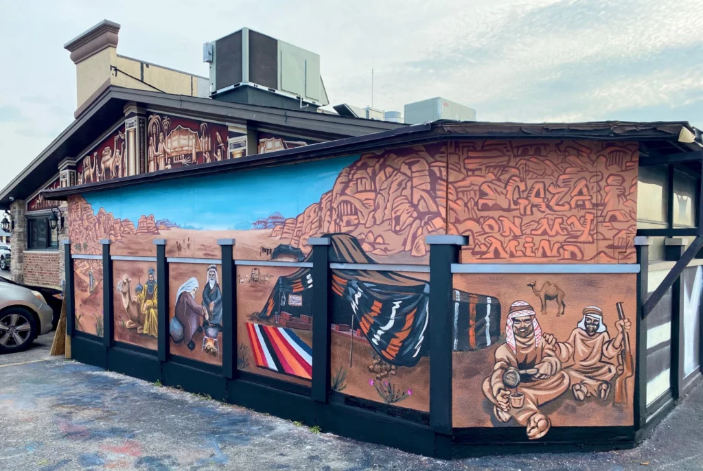 Gaza on My Mind, mural by Caleb Wassell: the full wrap-around restaurant wall painted with red desert cliffs, camels, a Bedouin tent and rug, an elderly couple cooking, and two men sharing coffee beneath the lettering Gaza on My Mind.