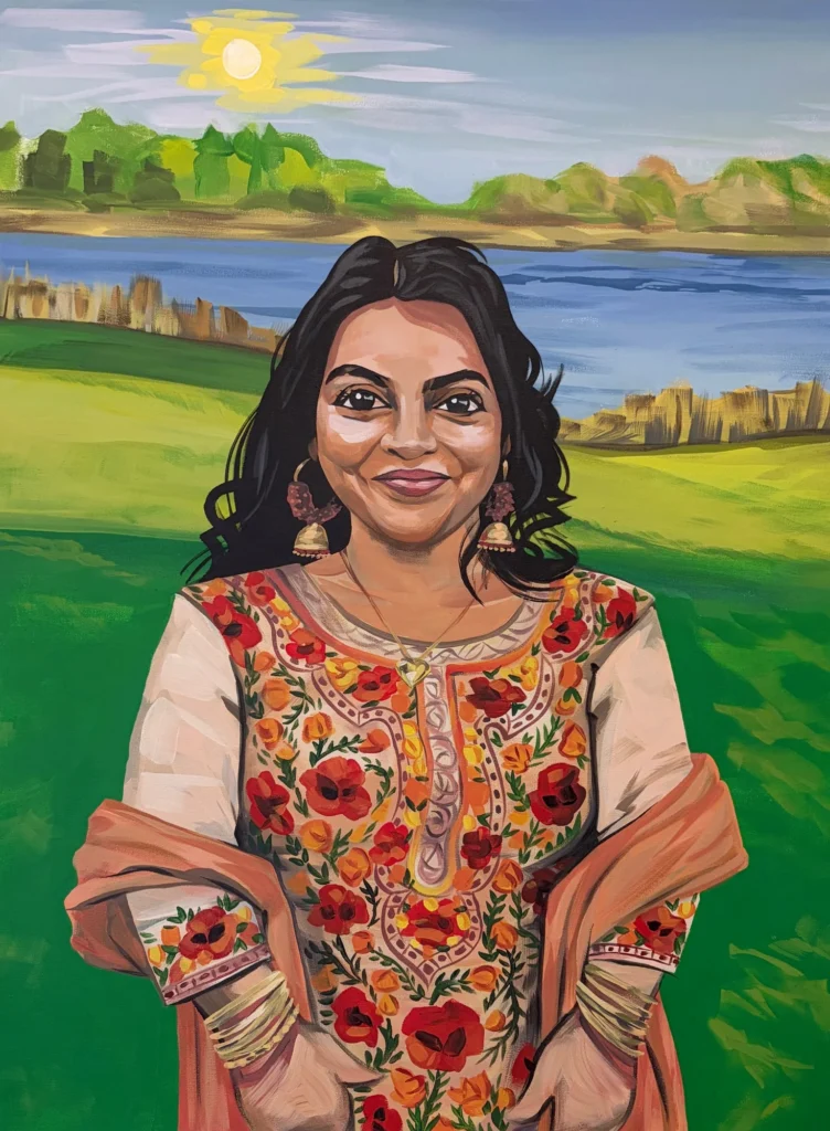 Portrait by the Lake, portrait by Caleb Wassell: a smiling woman in a floral embroidered kurta, gold jhumka earrings and a peach shawl stands on green grass beside a lake under a bright sun.